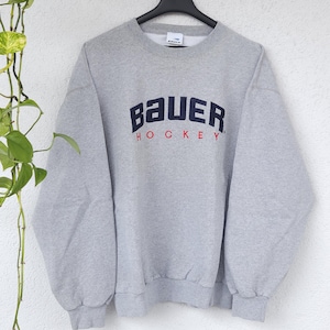 Könnte beinhalten: Graues Sweatshirt mit dem Wort "Bauer" in marineblauen Großbuchstaben über dem Wort "HOCKEY" in Rot. Das Sweatshirt hängt an einem schwarzen Kleiderbügel.