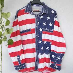 Könnte beinhalten: Ein Hemd mit Knopfleiste und patriotischem Design, das die amerikanische Flagge zeigt. Das Hemd hat rote und weiße Streifen auf der einen Seite und ein blaues Feld mit weißen Sternen auf der anderen Seite. Kragen, Manschetten und Taschen sind ebenfalls blau mit weißen Sternen.