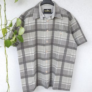 Könnte beinhalten: Kurzärmeliges Hemd mit grauem und weißem Karomuster. Das Hemd hat einen Kragen und eine Brusttasche mit dem gestickten Text "uncle sam". Es ist zugeknöpft und hängt an einem schwarzen Kleiderbügel.