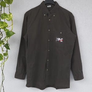 Könnte beinhalten: Ein dunkel olivgrünes Button-Down-Hemd mit langen Ärmeln, das an einem Kleiderbügel hängt. Das Hemd hat einen kleinen gestickten Aufnäher auf der Brust mit einem Logo in Rot, Weiß und Schwarz. Das Hemd ist aus strapazierfähigem Stoff.