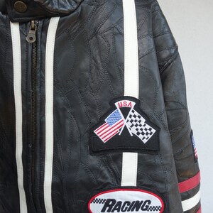 K&ouml;nnte beinhalten: Schwarze Lederjacke mit wei&szlig;en Rennstreifen und roten Akzenten. Die Jacke hat ein "Diamond Plate Genuine"-Etikett, einen "USA"-Patch mit gekreuzten amerikanischen und karierten Flaggen sowie einen "Racing"-Patch.