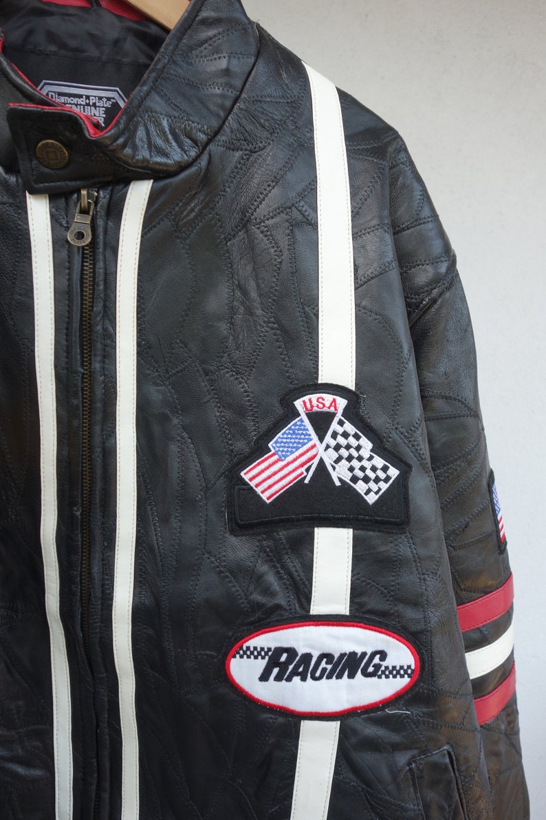 K&ouml;nnte beinhalten: Schwarze Lederjacke mit wei&szlig;en Rennstreifen und roten Akzenten. Die Jacke hat ein "Diamond Plate Genuine"-Etikett, einen "USA"-Patch mit gekreuzten amerikanischen und karierten Flaggen sowie einen "Racing"-Patch.