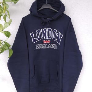 Könnte beinhalten: Marineblauer Kapuzenpullover mit dem Wort "LONDON" in weißen Blockbuchstaben, umrandet in Marineblau. Darunter befindet sich eine kleine Union Jack-Flagge und das Wort "ENGLAND". Der Hoodie hat eine Vordertasche und eine Kapuze mit Kordelzug.