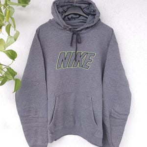 Op de afbeelding: Een grijze Nike hoodie met een voorzak en een capuchon met trekkoord. Het woord "NIKE" is op de borst gedrukt met een neon groene omlijning. De hoodie hangt aan een metalen hanger.