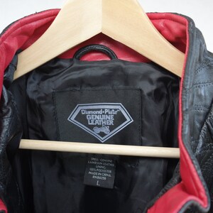 K&ouml;nnte beinhalten: Schwarze Lederjacke mit rotem Futter. Die Jacke hat ein rautenf&ouml;rmiges Etikett mit der Aufschrift "Diamond Plate Genuine Leather". Das Etikett enth&auml;lt auch den Text "Shell: Genuine Lambskin Leather, Lining: 100% Polyester, Made in China, RN 84256, L".