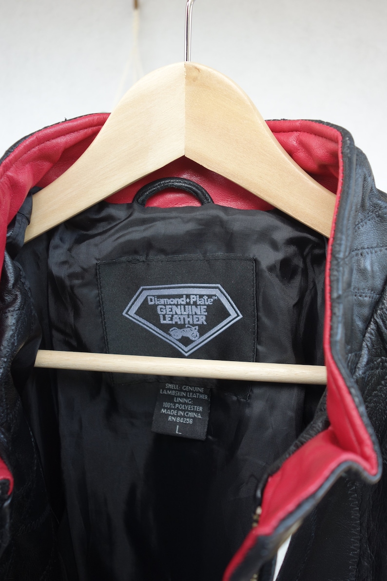 K&ouml;nnte beinhalten: Schwarze Lederjacke mit rotem Futter. Die Jacke hat ein rautenf&ouml;rmiges Etikett mit der Aufschrift "Diamond Plate Genuine Leather". Das Etikett enth&auml;lt auch den Text "Shell: Genuine Lambskin Leather, Lining: 100% Polyester, Made in China, RN 84256, L".