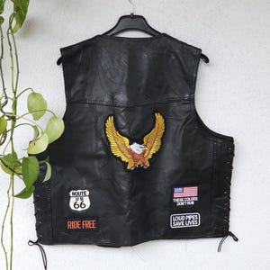 Könnte beinhalten: Schwarze Lederweste mit einem gestickten goldenen Adler-Patch. Mit Route 66 und amerikanischen Flaggen-Patches, mit Texten "RIDE FREE" und "THESE COLORS DON'T RUN, LOUD PIPES SAVE LIVES". Die Weste hat seitliche Schnürung.