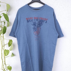 Peut inclure: T-shirt bleu à manches courtes avec l'inscription "BASS PRO SHOPS" en rouge et bleu. Le t-shirt présente un aigle et un écusson. Le texte "American Tradition Since 1972" est également visible.