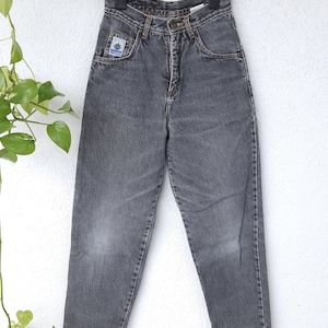 Può includere: Un paio di jeans grigi appesi a una gruccia nera. I jeans hanno vita alta, chiusura con bottone e cerniera e tasche. Una piccola toppa con un logo è visibile sulla tasca anteriore.