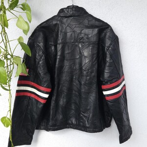 K&ouml;nnte beinhalten: Schwarze Lederjacke mit roten und wei&szlig;en Streifen an den &Auml;rmeln. Die Jacke hat einen Rei&szlig;verschluss und einen Stehkragen.
