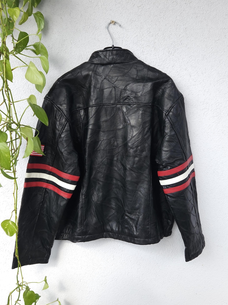 K&ouml;nnte beinhalten: Schwarze Lederjacke mit roten und wei&szlig;en Streifen an den &Auml;rmeln. Die Jacke hat einen Rei&szlig;verschluss und einen Stehkragen.