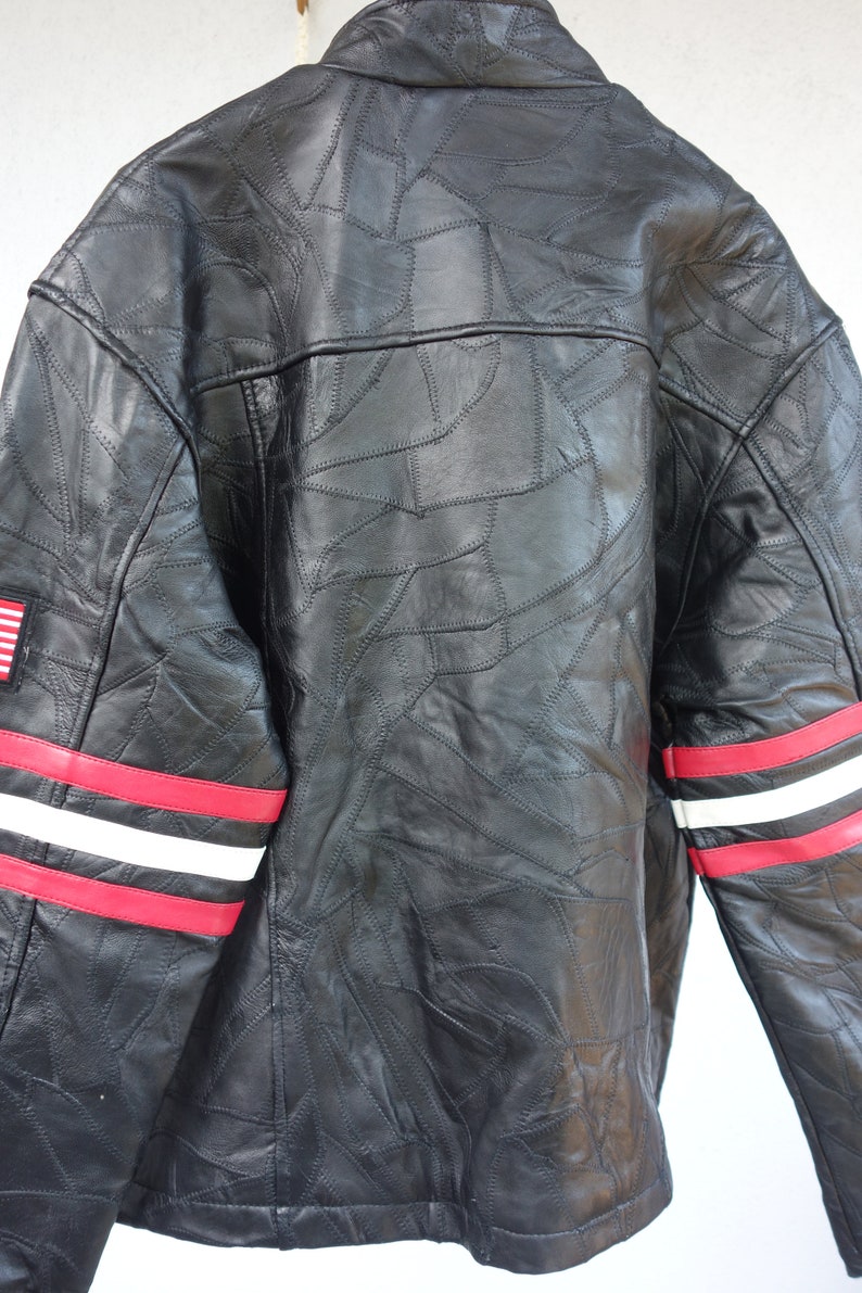 K&ouml;nnte beinhalten: Schwarze Lederjacke mit roten und wei&szlig;en Streifen an den &Auml;rmeln und einem kleinen amerikanischen Flaggen-Patch auf der linken Brust. Die Jacke ist aus Patchwork-Leder mit einem einzigartigen Design.