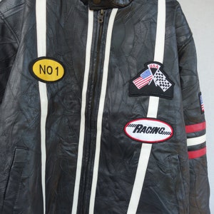 K&ouml;nnte beinhalten: Schwarze Leder-Rennjacke mit wei&szlig;en Streifen und roten Akzenten. Sie hat Patches mit dem Text "NO 1", "USA" und "RACING".