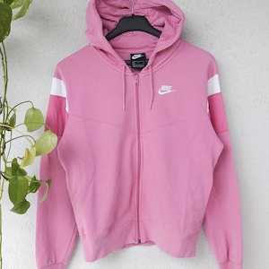 Könnte beinhalten: Rosa Kapuzenjacke mit Reißverschluss und weißen Akzenten an den Ärmeln. Das Nike-Logo ist auf der Brust zu sehen. Die Jacke hängt an einem schwarzen Kleiderbügel vor einer weißen Wand.