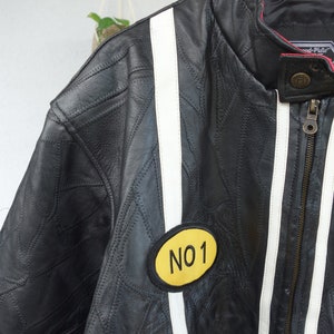 K&ouml;nnte beinhalten: Schwarze Lederjacke mit wei&szlig;en und roten Rennstreifen. Die Jacke hat einen gelben Aufn&auml;her mit dem Text "NO 1" auf der linken Seite.
