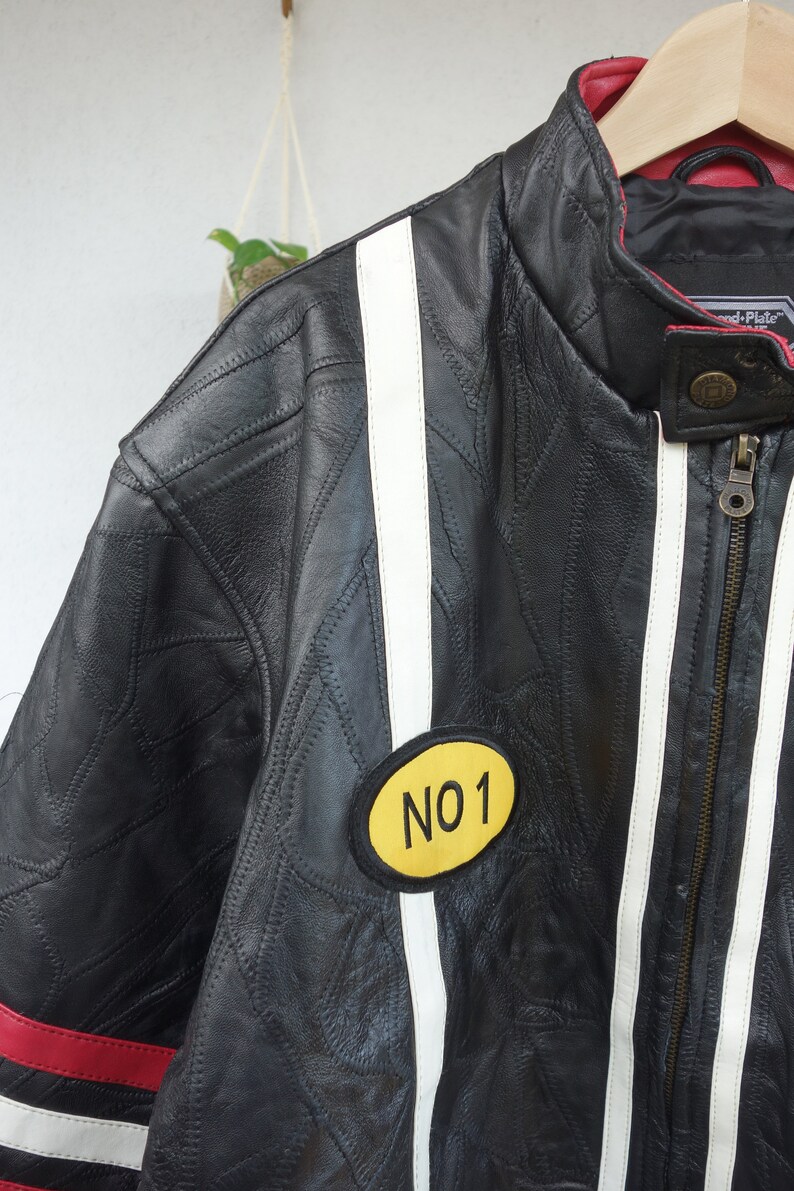 K&ouml;nnte beinhalten: Schwarze Lederjacke mit wei&szlig;en und roten Rennstreifen. Die Jacke hat einen gelben Aufn&auml;her mit dem Text "NO 1" auf der linken Seite.