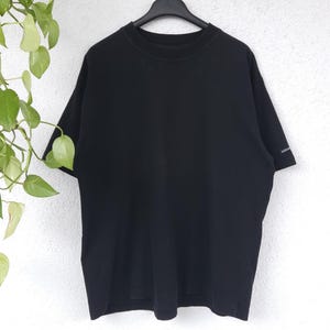 Könnte beinhalten: Einfarbiges schwarzes T-Shirt mit kurzen Ärmeln, hängt an einem schwarzen Kleiderbügel. Es hat einen Rundhalsausschnitt und ein kleines Logo am linken Ärmel. Der Hintergrund ist eine weiße Wand mit grünen Pflanzen.