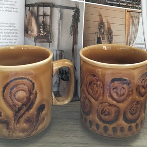 Può includere: Due tazze in ceramica marrone con motivi decorativi. Una tazza presenta un design a spirale, mentre l'altra ha un motivo circolare ripetuto. Entrambe le tazze hanno un manico e un interno marrone chiaro.