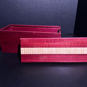 May include: A handcrafted wooden box with a deep purple hue. The lid features a light-colored stripe across the center. The box is open, revealing its interior. The wood grain is visible, adding texture to the design.