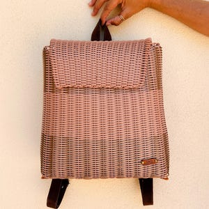 May include: A woven backpack in a light pink hue, featuring a flap closure and adjustable brown straps. The bag has a rectangular shape with a textured surface, and a small leather tag is attached. The bag is held by a hand.