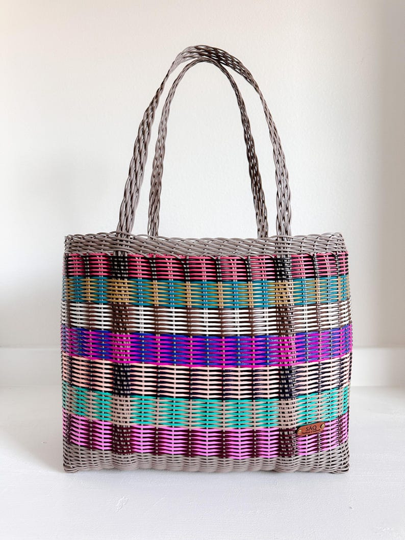 May include: A colourful woven tote bag with a rectangular shape and two handles. The bag has a striped pattern in pink, purple, teal, and brown, with a grey border and handles. A small brown tag is attached to the side.