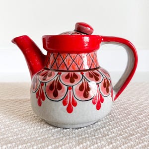 Può includere: Una teiera in ceramica rossa e bianca con un design dipinto a mano. La teiera ha un corpo rotondo e un beccuccio su un lato. Il manico è sul lato opposto al beccuccio. La teiera è decorata con un motivo rosso, bianco e marrone.