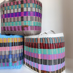 May include: Three woven storage baskets in various colours. The baskets are rectangular with handles and are made of woven plastic strips. The colours include grey, blue, white, pink, purple, teal, and black. Each basket has a small brown tag.
