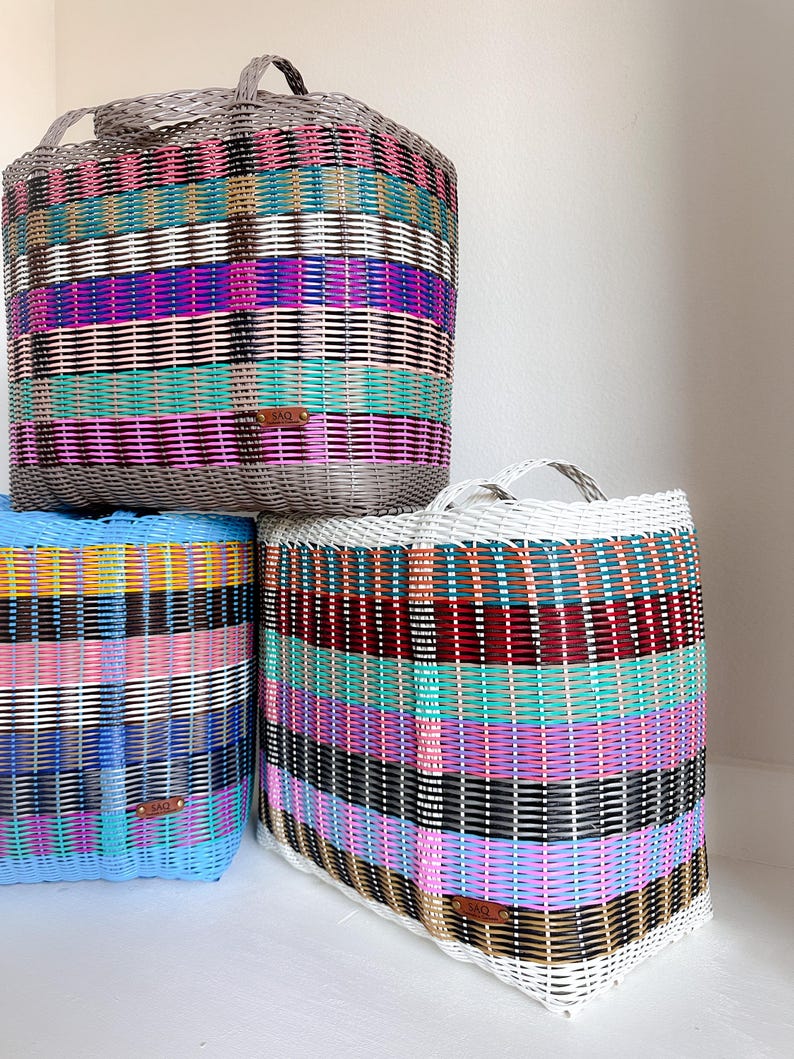May include: Three woven storage baskets in various colours. The baskets are rectangular with handles and are made of woven plastic strips. The colours include grey, blue, white, pink, purple, teal, and black. Each basket has a small brown tag.