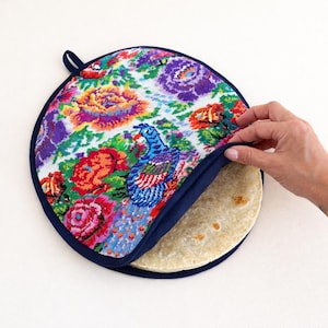 May include: A round, floral-patterned tortilla warmer with a navy blue trim. The fabric features a colorful peacock and flower design. The warmer holds several tortillas. A loop is attached for hanging.