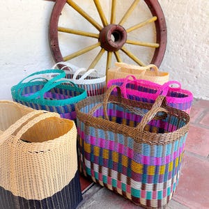 May include: A selection of brightly coloured woven baskets with handles. The baskets feature patterns in teal, pink, purple, and natural hues. A wooden cartwheel is visible in the background.