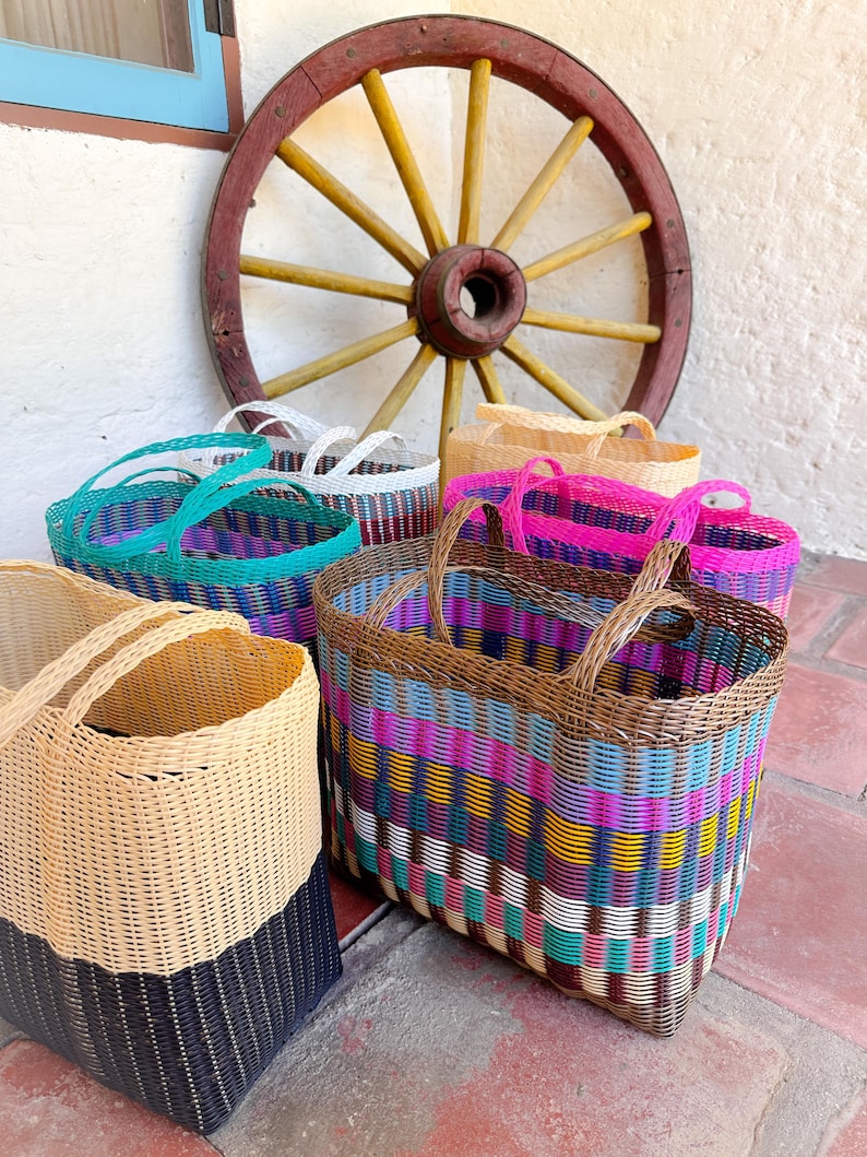 May include: A selection of brightly coloured woven baskets with handles. The baskets feature patterns in teal, pink, purple, and natural hues. A wooden cartwheel is visible in the background.
