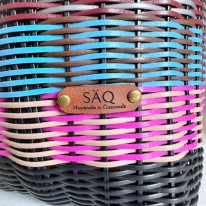 May include: Close-up of a woven basket with a colourful striped pattern. The basket features bands of black, pink, tan, blue, brown, grey, and burgundy. A leather tag reads "S&Auml;Q Handmade in Guatemala".