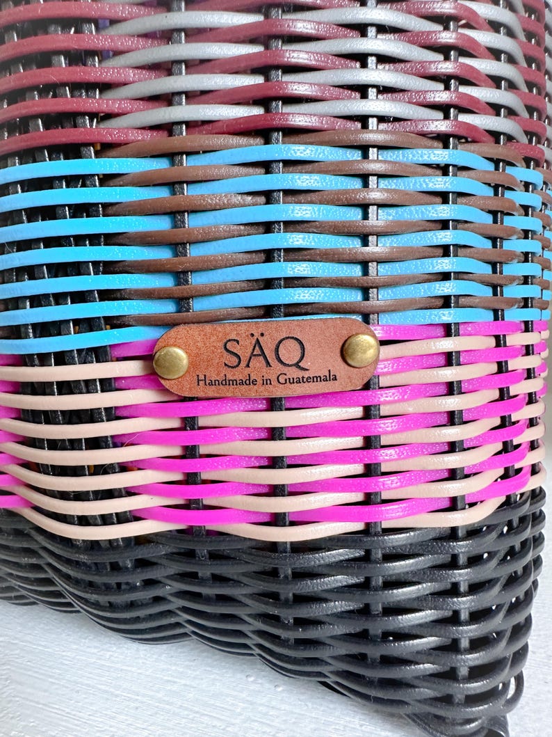 May include: Close-up of a woven basket with a colourful striped pattern. The basket features bands of black, pink, tan, blue, brown, grey, and burgundy. A leather tag reads "S&Auml;Q Handmade in Guatemala".