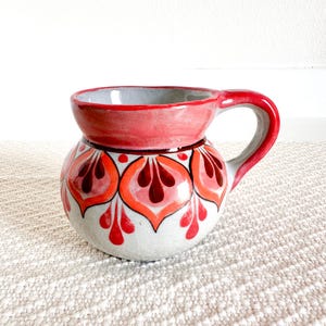Puede incluir: Una taza de cerámica roja y blanca con un diseño floral. La taza tiene un cuerpo redondeado y un asa.