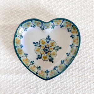 Puede incluir: Un plato de cerámica en forma de corazón con fondo blanco y un diseño floral verde y amarillo. El plato tiene un borde festoneado y un borde elevado.