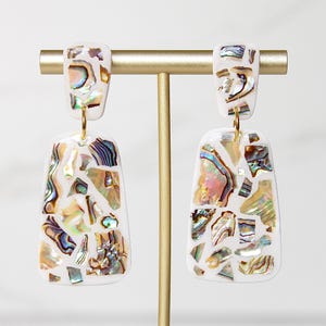 May include: A pair of white earrings with a rectangular shape, adorned with iridescent abalone shell fragments. The earrings hang from a gold-colored metal post. The abalone shell pieces display a mix of colors, including blue, green, and gold.