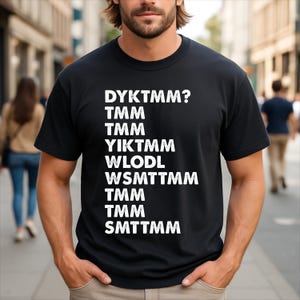 May include: A black short-sleeved t-shirt featuring white distressed-style text printed on the front. The text reads: DYKTMM? TMM TMM YIKTMM WLODL WSMTTMM TMM TMM SMTTMM. The shirt has a crew neckline and a standard fit, providing a simple and clean look.