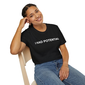 May include: Black t-shirt with the white text "I HAD POTENTIAL". The shirt is paired with blue jeans. A person is wearing the shirt and sitting on a wooden chair.