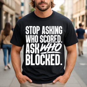 May include: A black short-sleeved t-shirt featuring bold white text that reads: STOP ASKING WHO SCORED. ASK WHO BLOCKED. The lettering is arranged in a clean, block-style font with the word WHO stylised in a brush-stroke effect across the chest.
