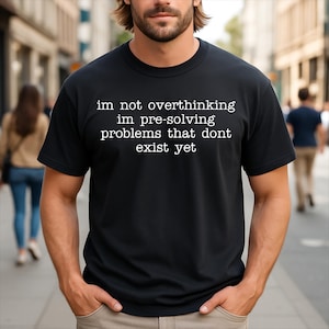 May include: A black short-sleeved t-shirt featuring white text that reads: im not overthinking im pre-solving problems that dont exist yet. The shirt has a crew neckline and a standard fit, providing a clear view of the printed message on the chest.