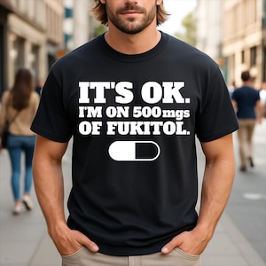 May include: A black short-sleeved t-shirt featuring white text that reads IT'S OK. I'M ON 500 mgs OF FUKITOL. Below the text is a graphic of a pill capsule that is half white and half black. The shirt has a crew neckline and a standard fit.