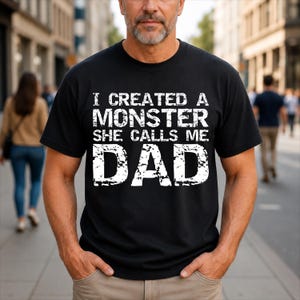 May include: A black short-sleeved t-shirt featuring white distressed-style text that reads I CREATED A MONSTER SHE CALLS ME DAD. The graphic is centred on the chest and uses a bold, blocky font with a weathered, textured appearance throughout.