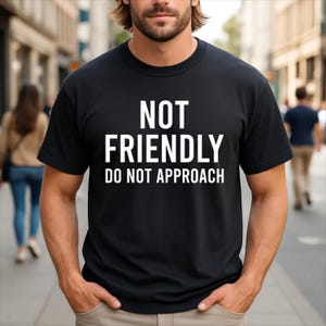 Op de afbeelding: Een zwart T-shirt met korte mouwen en de witte tekst NOT FRIENDLY DO NOT APPROACH in een vet, schreefloos lettertype op de borst gedrukt. Het kledingstuk heeft een klassieke ronde hals en een rechte zoom, wat zorgt voor een strak ontwerp.