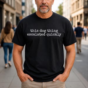 Puede incluir: Una camiseta negra de manga corta con el texto blanco "this dog thing escalated quickly" impreso en una fuente redondeada e informal en el pecho. La prenda tiene un cuello redondo estándar y un dobladillo recto, ofreciendo un diseño sencillo.