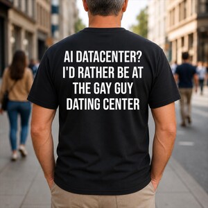 Op de afbeelding: Een zwart T-shirt met korte mouwen en witte tekst op de achterkant: AI DATACENTER? I'D RATHER BE AT THE GAY GUY DATING CENTER. Het shirt heeft een ronde hals en wordt gedragen boven een lichte broek in een casual, alledaagse stijl.