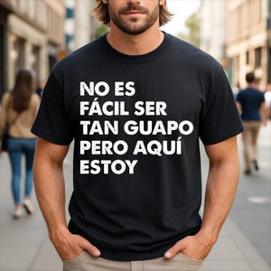 May include: A black short-sleeved t-shirt featuring white text printed on the chest that reads: NO ES FÁCIL SER TAN GUAPO PERO AQUÍ ESTOY. The garment has a crew neckline and a standard fit, displaying the bold lettering clearly against the dark fabric.