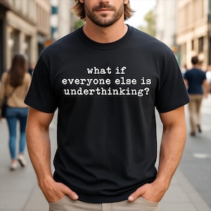 Pode incluir: Uma t-shirt preta de manga curta com texto branco impresso no peito que diz: what if everyone else is underthinking? A peça tem um decote redondo clássico e um corte padrão, proporcionando uma aparência limpa e simples.
