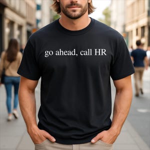 May include: A black short-sleeved t-shirt featuring the white text "go ahead, call HR" printed across the chest. The garment has a classic crew neckline and a standard fit, constructed from a solid black fabric with clean white lettering.