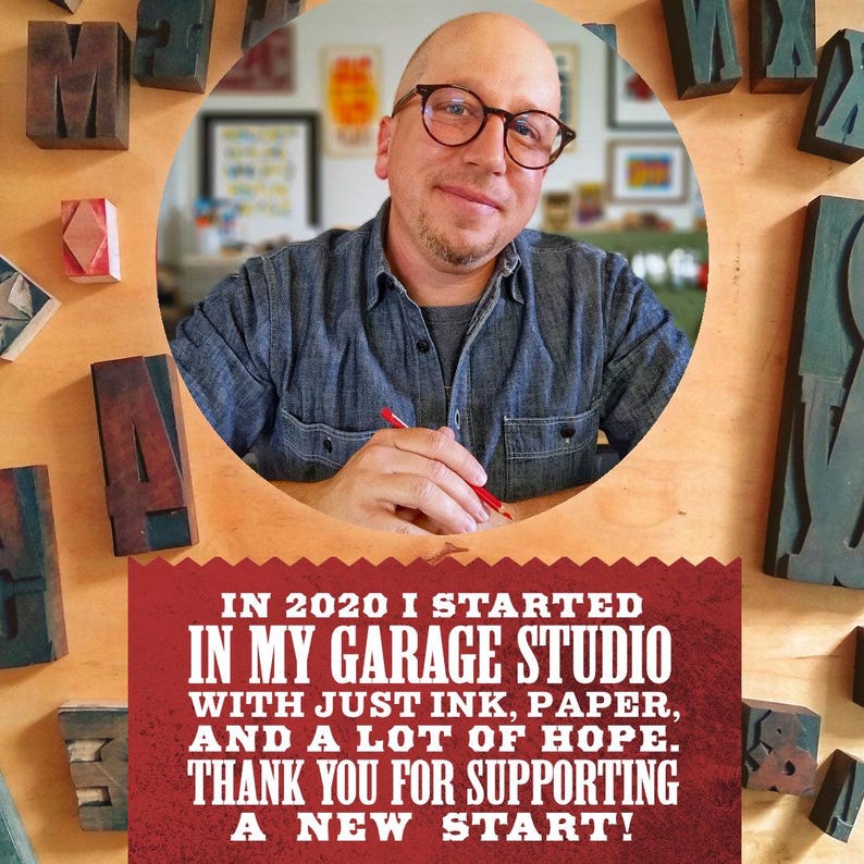 A man sitting at a desk in a garage studio, surrounded by various craft supplies and materials, with a sign that reads "In 2020 I started in my garage studio with just ink, paper, and a lot of hope. Thank you for supporting a new start!"