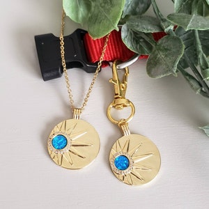 May include: A gold-toned necklace and a matching gold-toned pet tag, both featuring a circular pendant with a sunburst design, a central blue opal-like stone, and a halo of small clear crystals. The tag includes a gold-toned lobster clasp.